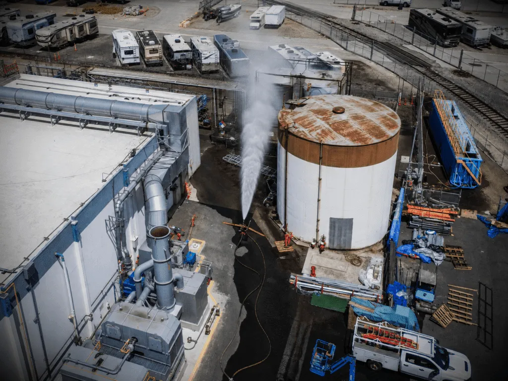 Industrial plant scene with a large cylindrical storage tank and steaming pipe system surrounding a gray building area.