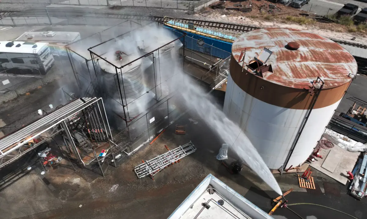 Industrial site with a high-pressure water jet spraying from a hose toward a large cylindrical storage tank, surrounded by scaffolding and equipment.