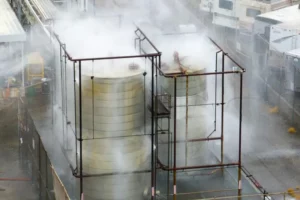 Industrial site with large cylindrical storage tanks surrounded by steam and piping fractures in a metal framework